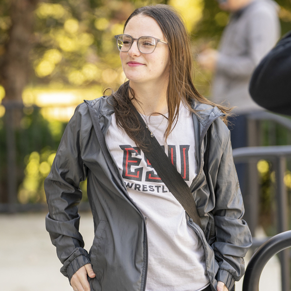 Student wearing a black jacket and glasses standing outdoors with trees in the background.