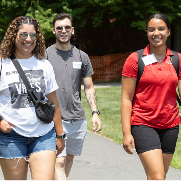 Three smiling students walk along a paved path on a sunny day, carrying backpacks.