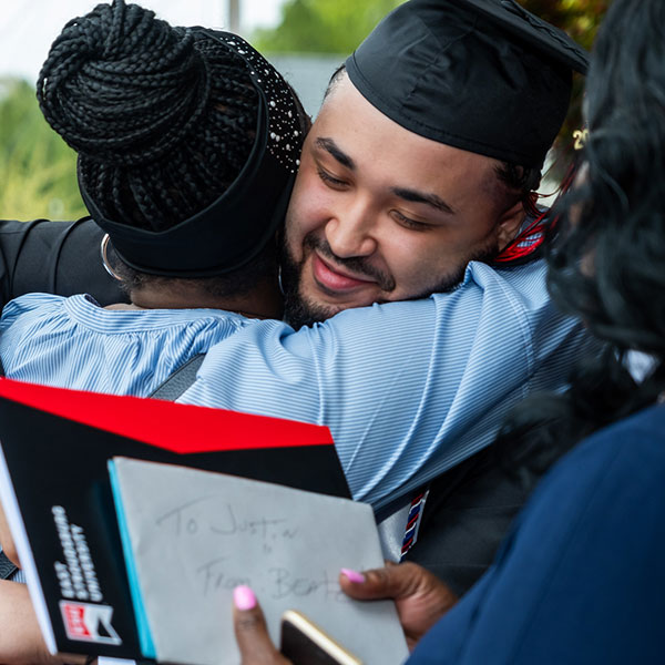 A graduate in a cap and gown hugging another person.