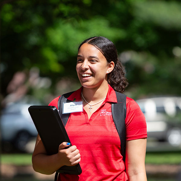 Student in a red polo smiling, holding a black folder, outdoors.