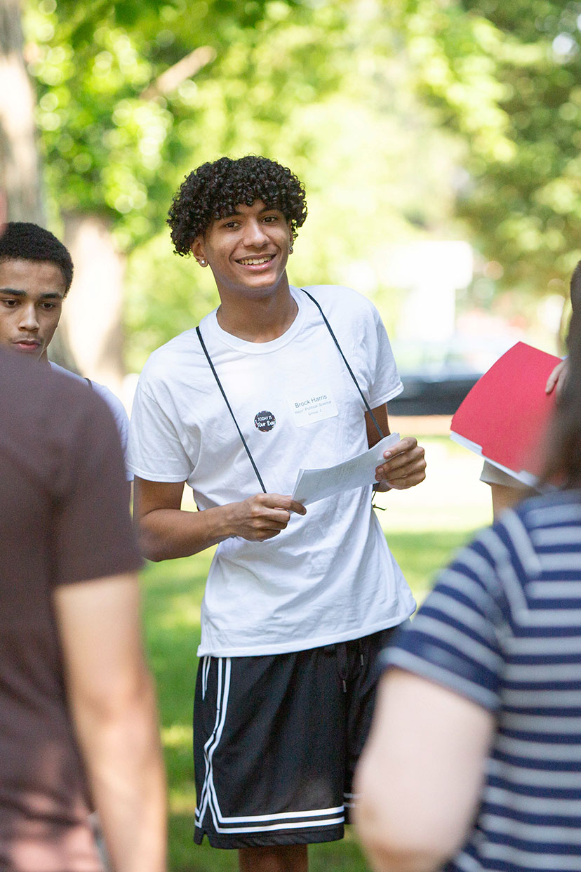 A group of students chatting outdoors, with a young man in the center holding a paper and smiling.