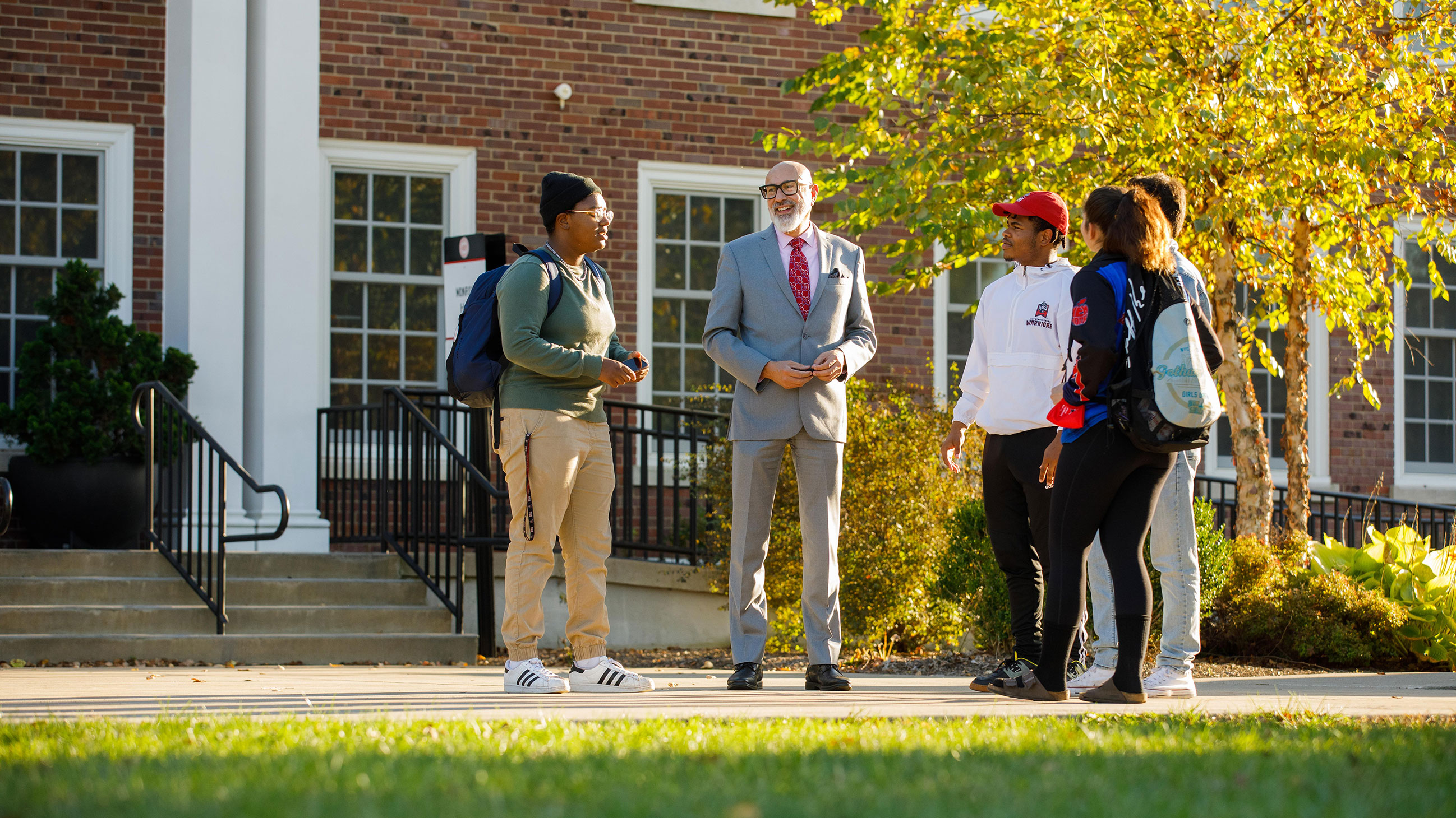 Five people standing and chatting outside a brick building on a sunny day.